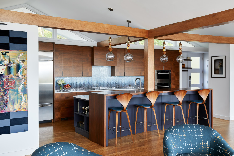 Midcentury galley open plan kitchen in New York with flat-panel cabinets, dark wood cabinets, stainless steel appliances, medium hardwood flooring, an island, brown floors, white worktops, exposed beams and a vaulted ceiling.