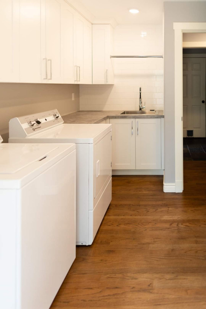 Photo of a large transitional l-shaped utility room in Seattle with a drop-in sink, shaker cabinets, white cabinets, laminate benchtops, white splashback, porcelain splashback, white walls, dark hardwood floors, a side-by-side washer and dryer, brown floor and grey benchtop.
