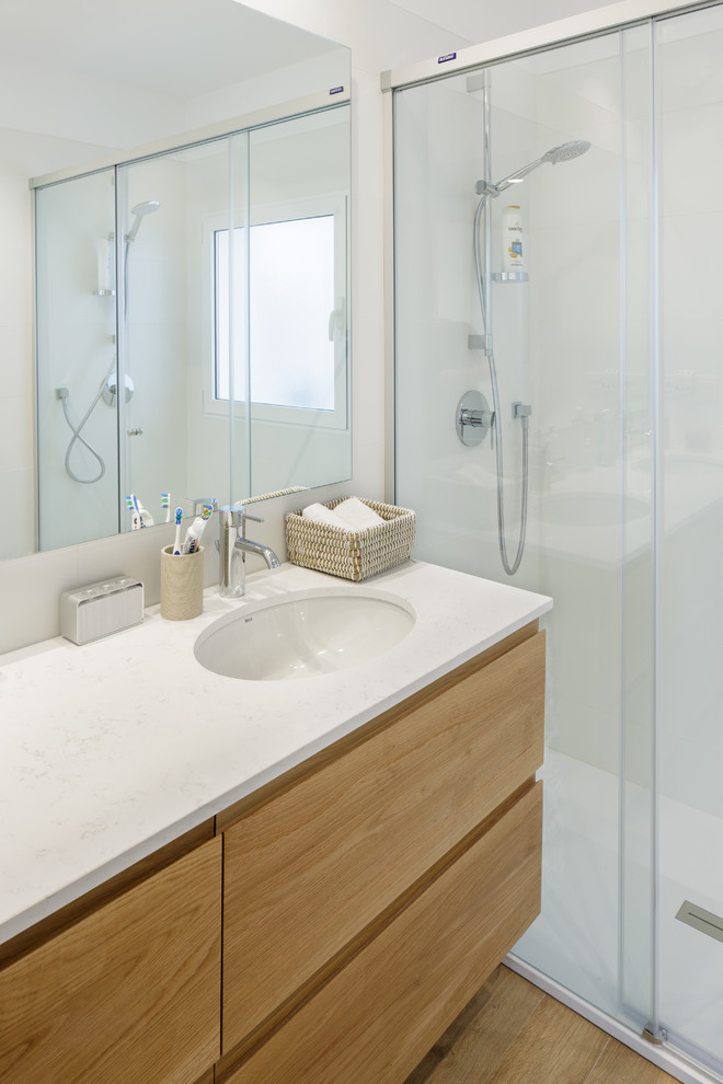 This is an example of a modern master wet room bathroom in Alicante-Costa Blanca with flat-panel cabinets, medium wood cabinets, a wall-mount toilet, ceramic tile, white walls, medium hardwood floors, an undermount sink, engineered quartz benchtops, a hinged shower door and white benchtops.