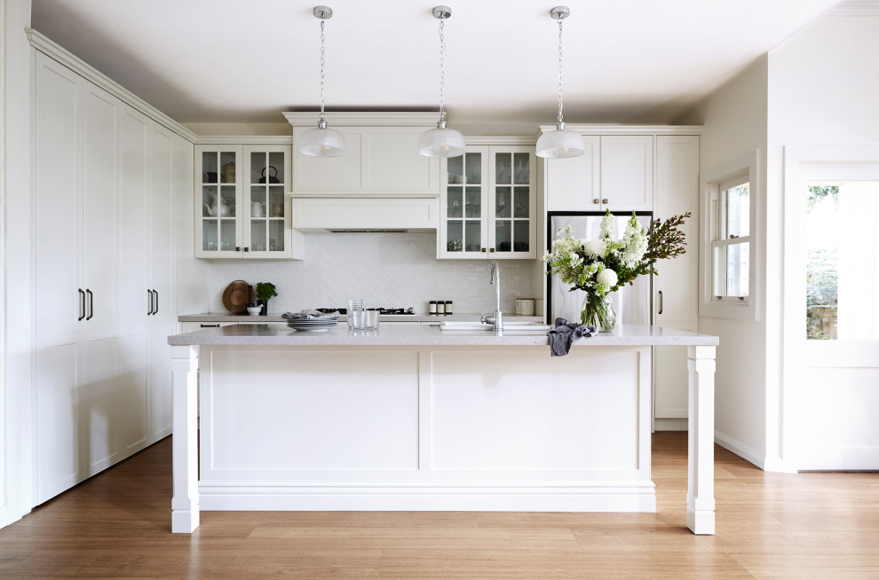 Mid-sized traditional l-shaped open plan kitchen in Sydney with a farmhouse sink, shaker cabinets, white cabinets, quartz benchtops, white splashback, porcelain splashback, coloured appliances, vinyl floors, with island, brown floor and grey benchtop.