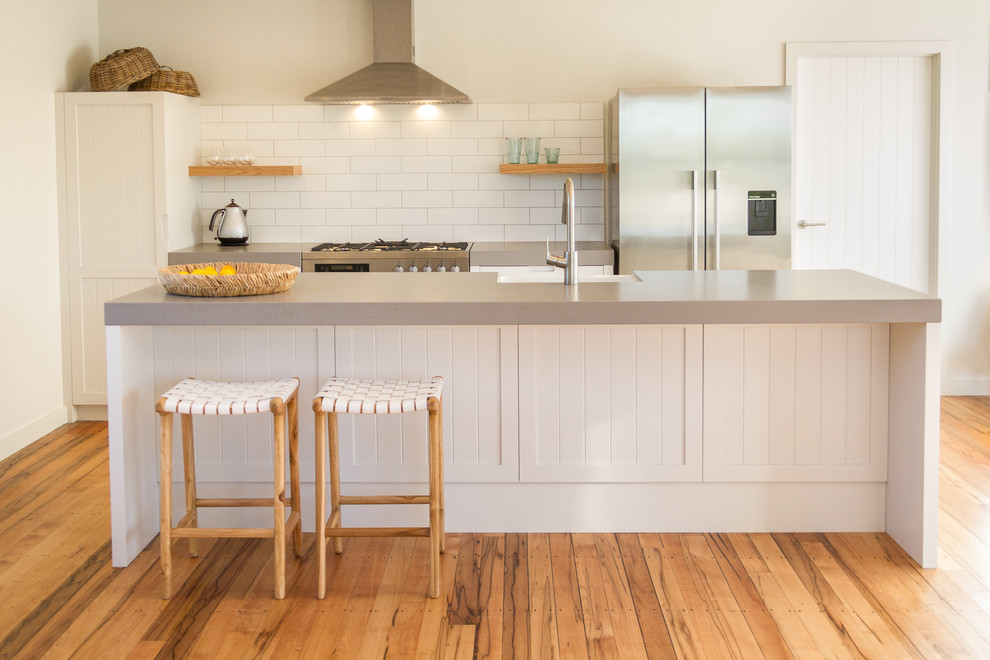 Inspiration for a small traditional galley open plan kitchen in Napier-Hastings with a farmhouse sink, recessed-panel cabinets, white cabinets, quartz benchtops, white splashback, ceramic splashback, stainless steel appliances, medium hardwood floors, with island, brown floor and grey benchtop.