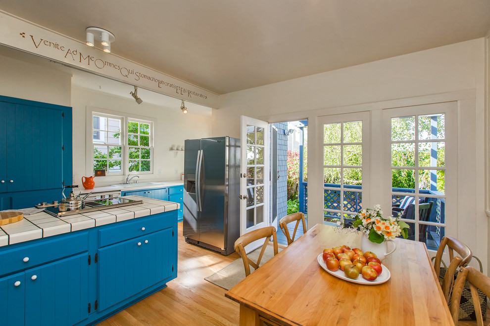 This is an example of an arts and crafts galley eat-in kitchen in San Francisco with louvered cabinets, blue cabinets, tile benchtops, stainless steel appliances, light hardwood floors and with island.