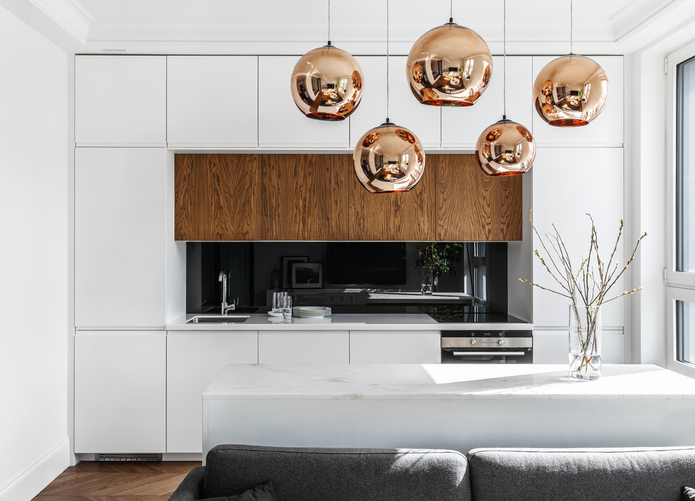 Photo of a contemporary single-wall open plan kitchen in Moscow with an undermount sink, flat-panel cabinets, white cabinets, black splashback, glass sheet splashback, medium hardwood floors, with island, brown floor, white benchtop and stainless steel appliances.