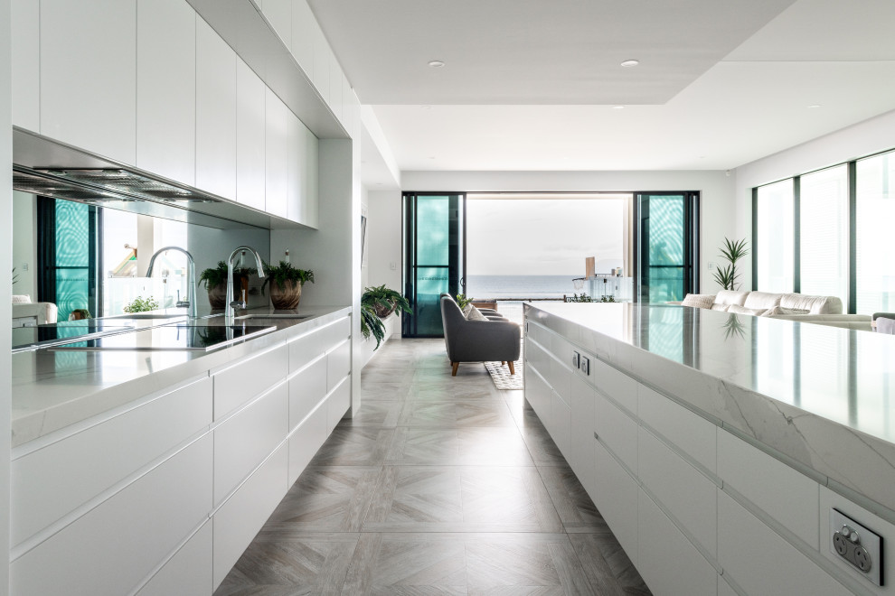Photo of a large beach style galley open plan kitchen in Gold Coast - Tweed with an undermount sink, flat-panel cabinets, white cabinets, quartz benchtops, mirror splashback, stainless steel appliances, porcelain floors, with island, multi-coloured floor and white benchtop.