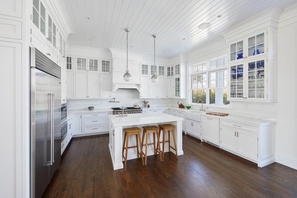 Photo of a large country u-shaped eat-in kitchen in New York with a farmhouse sink, beaded inset cabinets, white cabinets, quartzite benchtops, white splashback, stainless steel appliances, dark hardwood floors, with island, brown floor and white benchtop.