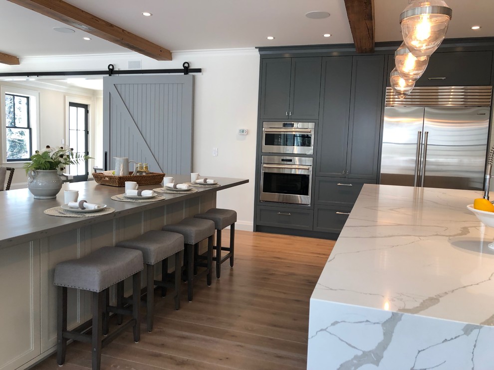 This is an example of an expansive country l-shaped kitchen in Boston with a single-bowl sink, recessed-panel cabinets, grey cabinets, quartz benchtops, white splashback, stone slab splashback, stainless steel appliances, light hardwood floors, multiple islands, brown floor and multi-coloured benchtop.