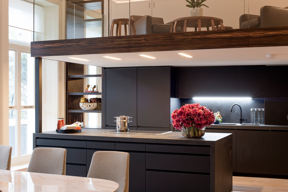Photo of an expansive midcentury galley open plan kitchen in London with a drop-in sink, glass-front cabinets, black cabinets, glass benchtops, black splashback, porcelain splashback, panelled appliances, with island and black benchtop.
