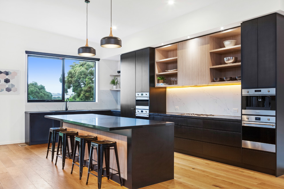 Design ideas for a contemporary l-shaped open plan kitchen in Melbourne with flat-panel cabinets, white splashback, black appliances, medium hardwood floors, with island and black benchtop.