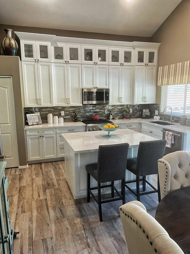 Mid-sized transitional l-shaped open plan kitchen in Phoenix with an undermount sink, shaker cabinets, white cabinets, quartz benchtops, multi-coloured splashback, glass sheet splashback, stainless steel appliances, vinyl floors, with island, beige floor and white benchtop.