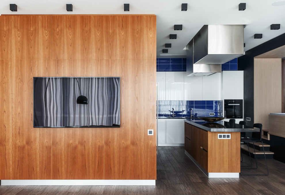 Expansive contemporary l-shaped open plan kitchen in Moscow with an undermount sink, flat-panel cabinets, white cabinets, granite benchtops, blue splashback, glass tile splashback, black appliances, medium hardwood floors, with island, brown floor and black benchtop.