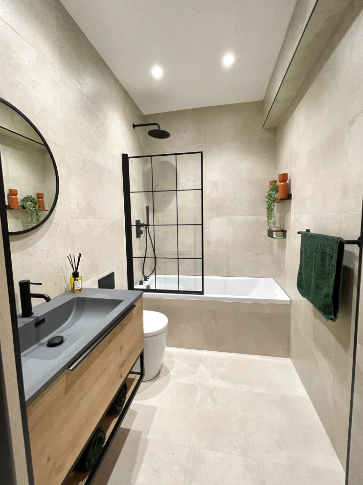 Photo of a mid-sized modern kids bathroom in Hertfordshire with flat-panel cabinets, medium wood cabinets, a drop-in tub, a shower/bathtub combo, a two-piece toilet, beige tile, porcelain tile, beige walls, porcelain floors, an integrated sink, beige floor, a hinged shower door, black benchtops, a single vanity and a floating vanity.