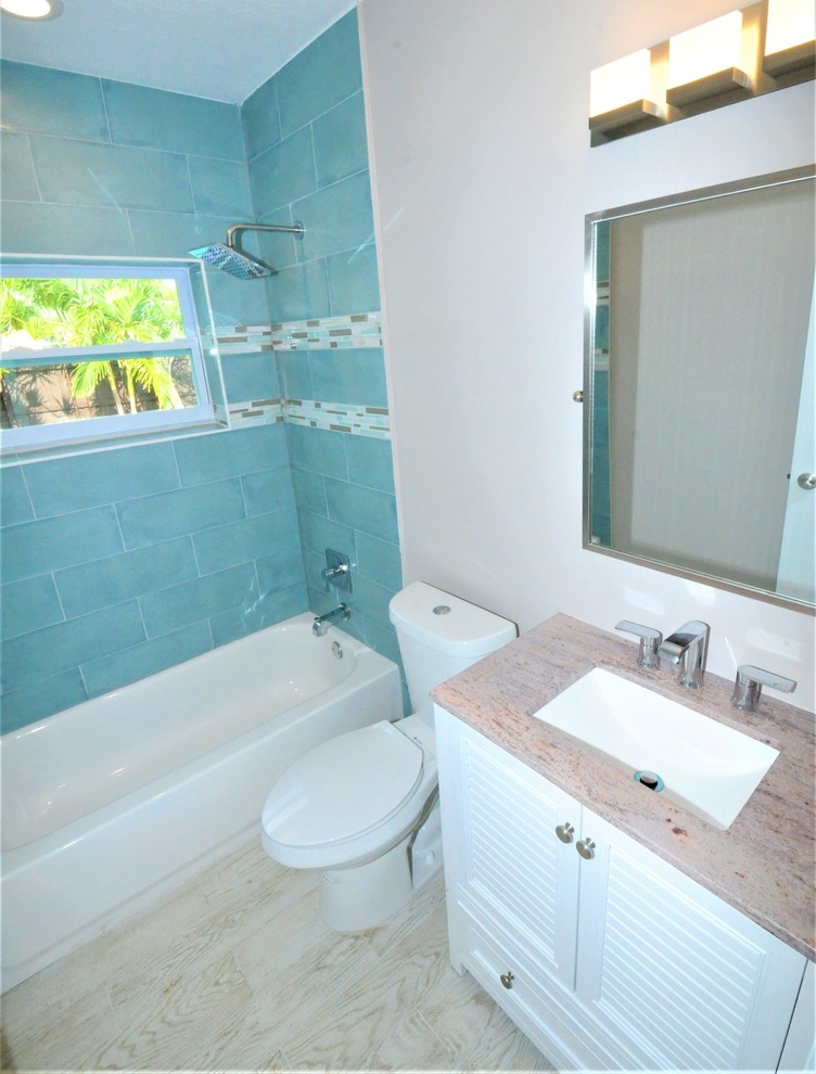 This is an example of a mid-sized contemporary kids bathroom in Miami with furniture-like cabinets, white cabinets, a drop-in tub, an alcove shower, a two-piece toilet, blue tile, ceramic tile, beige walls, ceramic floors, an undermount sink, granite benchtops, beige floor, a shower curtain and multi-coloured benchtops.