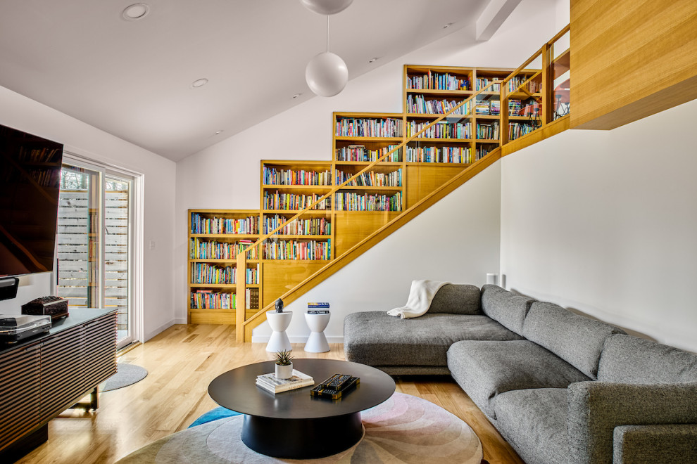 Cette image montre une grande salle de séjour vintage ouverte avec une bibliothèque ou un coin lecture, un mur blanc, un sol en bois brun, un téléviseur fixé au mur, un sol beige et un plafond voûté.
