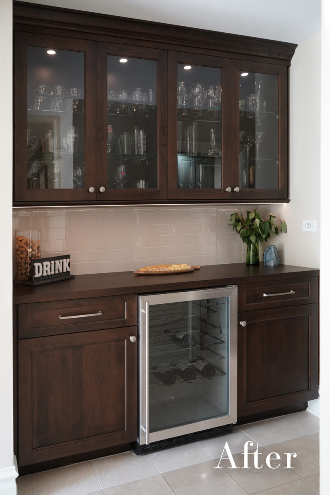 Photo of a large transitional single-wall home bar in Chicago with no sink, glass-front cabinets, dark wood cabinets, wood benchtops, beige splashback, ceramic splashback, ceramic floors, beige floor and brown benchtop.