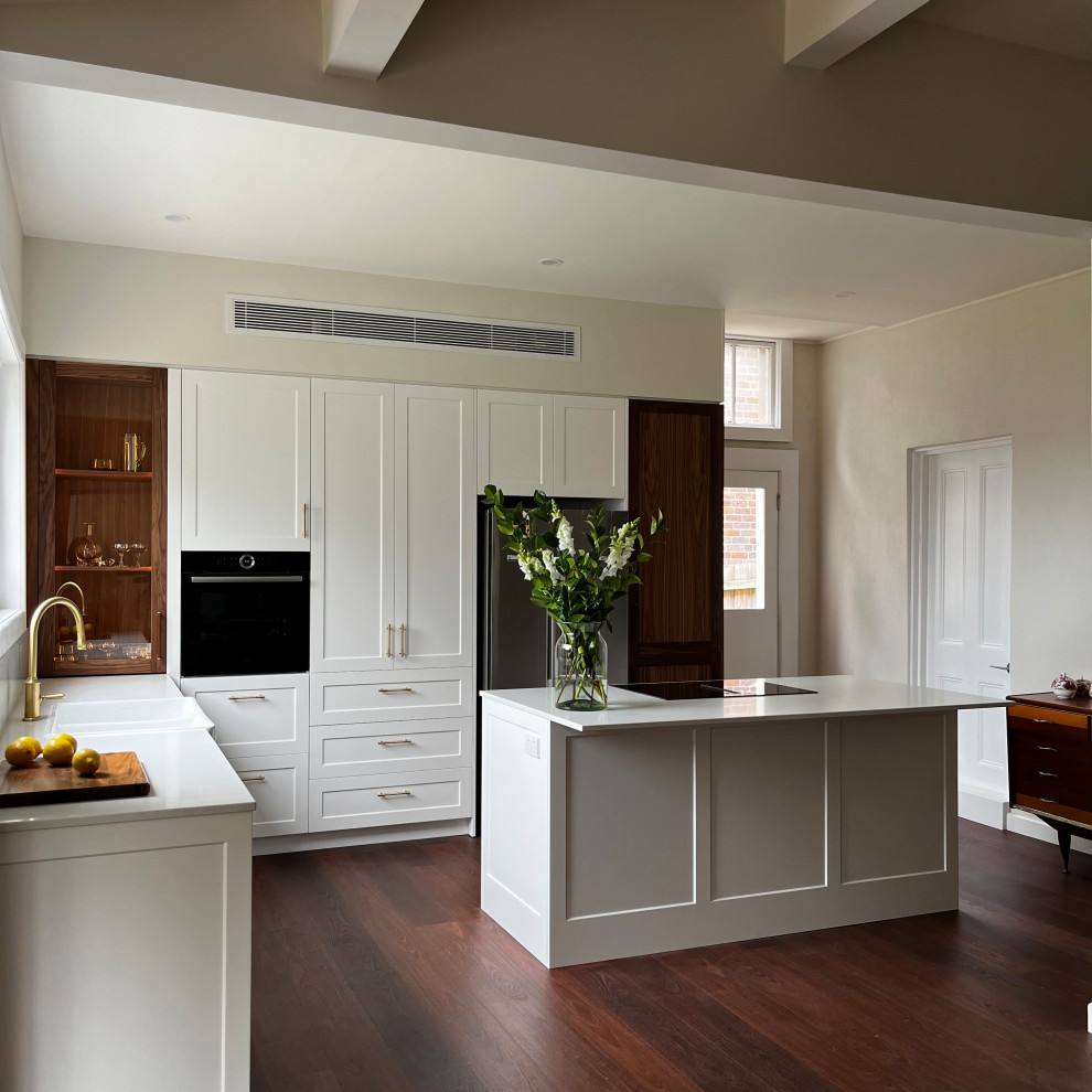 Photo of a mid-sized traditional l-shaped eat-in kitchen with a farmhouse sink, shaker cabinets, white cabinets, quartz benchtops, white splashback, engineered quartz splashback, black appliances, dark hardwood floors, with island, brown floor and white benchtop.