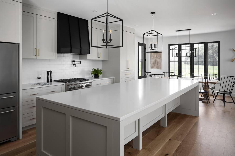 Large country single-wall eat-in kitchen in Toronto with a farmhouse sink, shaker cabinets, grey cabinets, quartz benchtops, white splashback, subway tile splashback, stainless steel appliances, light hardwood floors, with island, brown floor and grey benchtop.