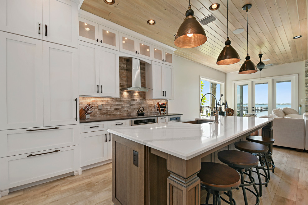 Photo of a large transitional l-shaped eat-in kitchen in Orlando with a farmhouse sink, shaker cabinets, white cabinets, beige splashback, subway tile splashback, stainless steel appliances, light hardwood floors, with island, marble benchtops and beige floor.
