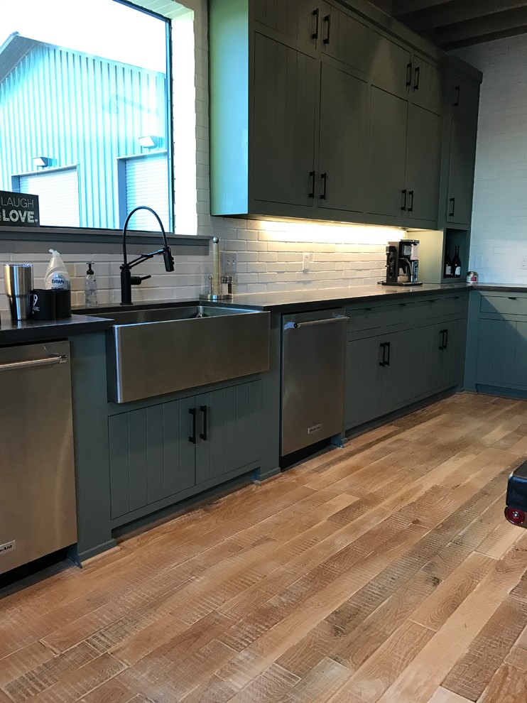 This is an example of an expansive industrial u-shaped open plan kitchen in Dallas with a farmhouse sink, green cabinets, concrete benchtops, white splashback, brick splashback, stainless steel appliances, light hardwood floors, with island, beige floor and flat-panel cabinets.