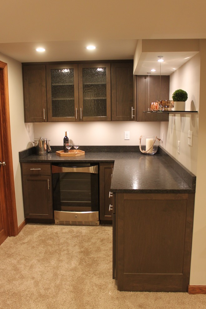 Photo of a small transitional l-shaped home bar in Milwaukee with no sink, shaker cabinets, dark wood cabinets, granite benchtops, brown splashback, stone slab splashback and carpet.