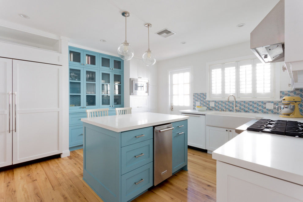Design ideas for a small coastal l-shaped enclosed kitchen in Los Angeles with a belfast sink, shaker cabinets, blue cabinets, composite countertops, blue splashback, ceramic splashback, stainless steel appliances, light hardwood flooring, an island, brown floors and white worktops.