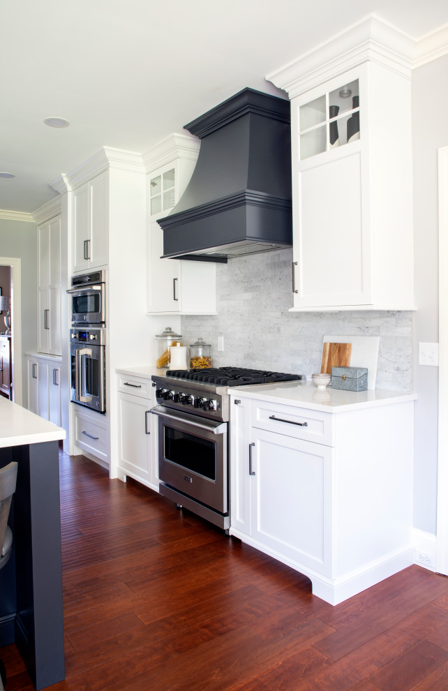 Example of a transitional medium tone wood floor kitchen design with an undermount sink, shaker cabinets, quartz countertops, gray backsplash, porcelain backsplash, stainless steel appliances, an island and white countertops