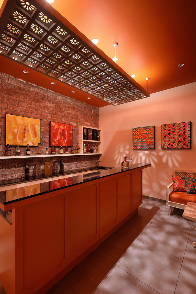 Design ideas for an asian galley wet bar in Ahmedabad with a drop-in sink, recessed-panel cabinets, orange cabinets, brown splashback, brick splashback, grey floor and black benchtop.