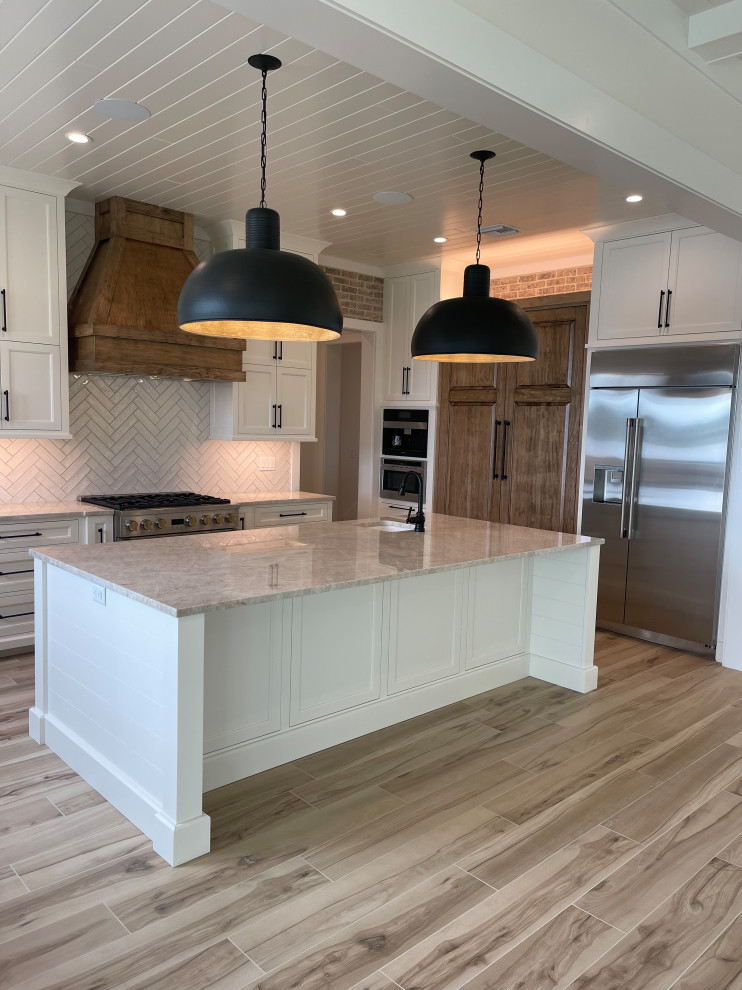 Photo of a mid-sized u-shaped open plan kitchen in Orlando with a farmhouse sink, shaker cabinets, white cabinets, quartzite benchtops, white splashback, brick splashback, stainless steel appliances, ceramic floors, with island, beige floor, beige benchtop and timber.