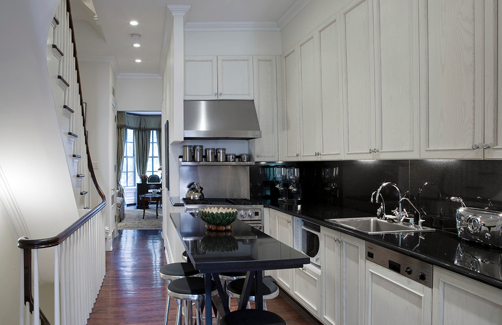 Photo of a traditional l-shaped kitchen in New York with a drop-in sink, recessed-panel cabinets, black splashback, glass sheet splashback, stainless steel appliances and dark hardwood floors.