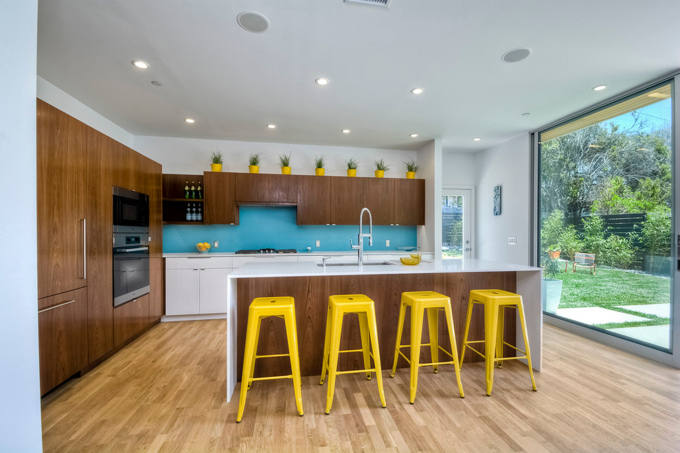 Design ideas for a midcentury galley kitchen in San Diego with an undermount sink, flat-panel cabinets, dark wood cabinets, blue splashback, glass sheet splashback, stainless steel appliances, light hardwood floors and with island.