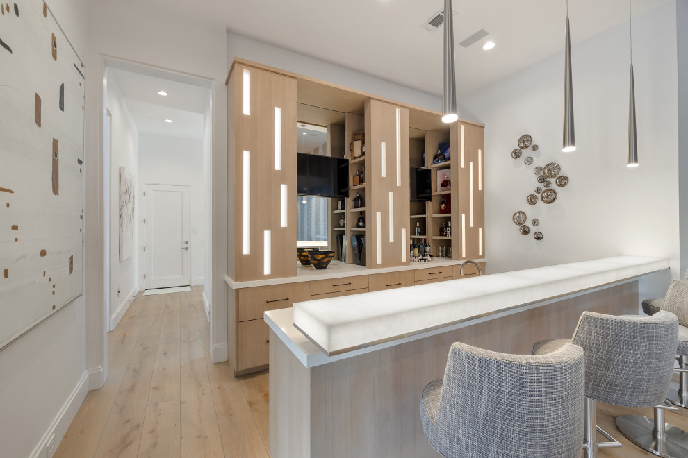 This is an example of a large contemporary galley seated home bar in Houston with an undermount sink, flat-panel cabinets, light wood cabinets, onyx benchtops, white splashback, mirror splashback, light hardwood floors and white benchtop.