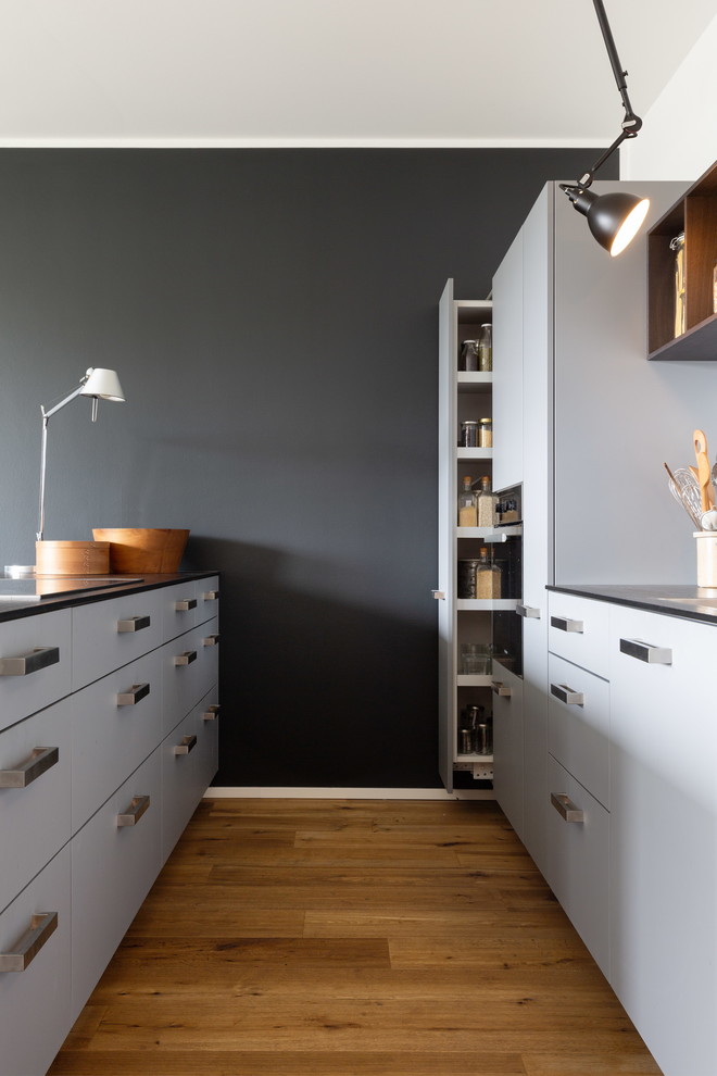 Inspiration for a mid-sized contemporary galley eat-in kitchen in Cologne with a single-bowl sink, flat-panel cabinets, grey cabinets, wood benchtops, white splashback, glass tile splashback, black appliances, dark hardwood floors, with island, brown floor and brown benchtop.
