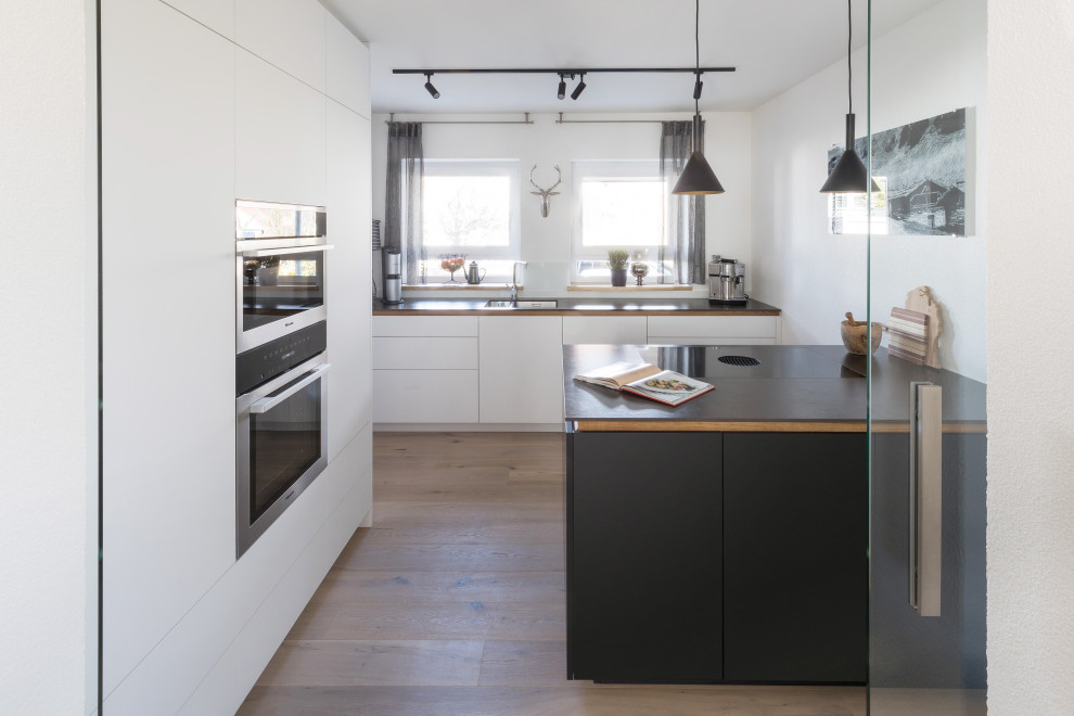 На фото: отдельная, параллельная кухня среднего размера в современном стиле с белыми фасадами, белым фартуком, светлым паркетным полом, островом и черной столешницей