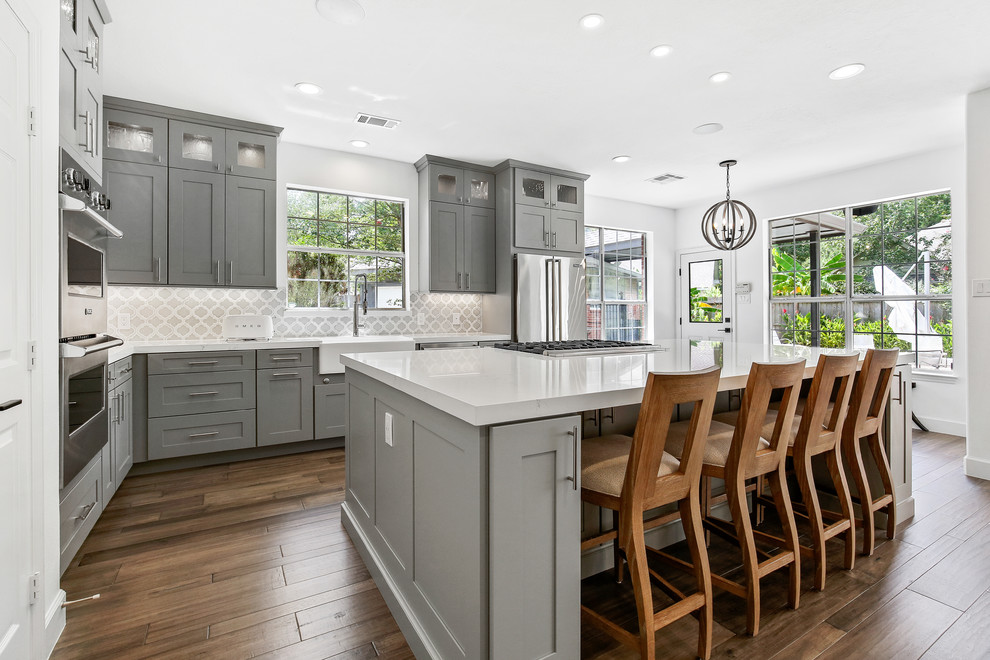 Design ideas for a mid-sized transitional l-shaped kitchen in Houston with a farmhouse sink, shaker cabinets, grey cabinets, quartzite benchtops, porcelain splashback, stainless steel appliances, with island, brown floor, white benchtop, multi-coloured splashback and dark hardwood floors.