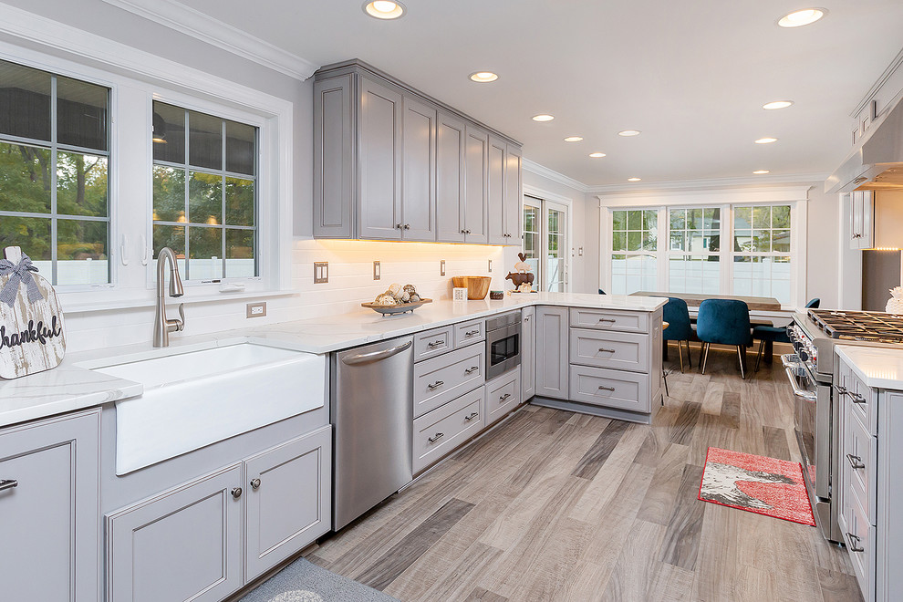 Photo of a large transitional u-shaped eat-in kitchen in Philadelphia with a farmhouse sink, raised-panel cabinets, grey cabinets, quartz benchtops, white splashback, ceramic splashback, stainless steel appliances, laminate floors, no island, brown floor and white benchtop.