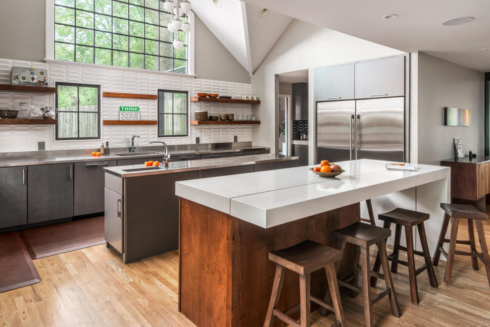 This is an example of a contemporary l-shaped kitchen in Nashville with flat-panel cabinets, grey cabinets, stainless steel benchtops, white splashback, stainless steel appliances, medium hardwood floors, multiple islands, brown floor, grey benchtop and vaulted.