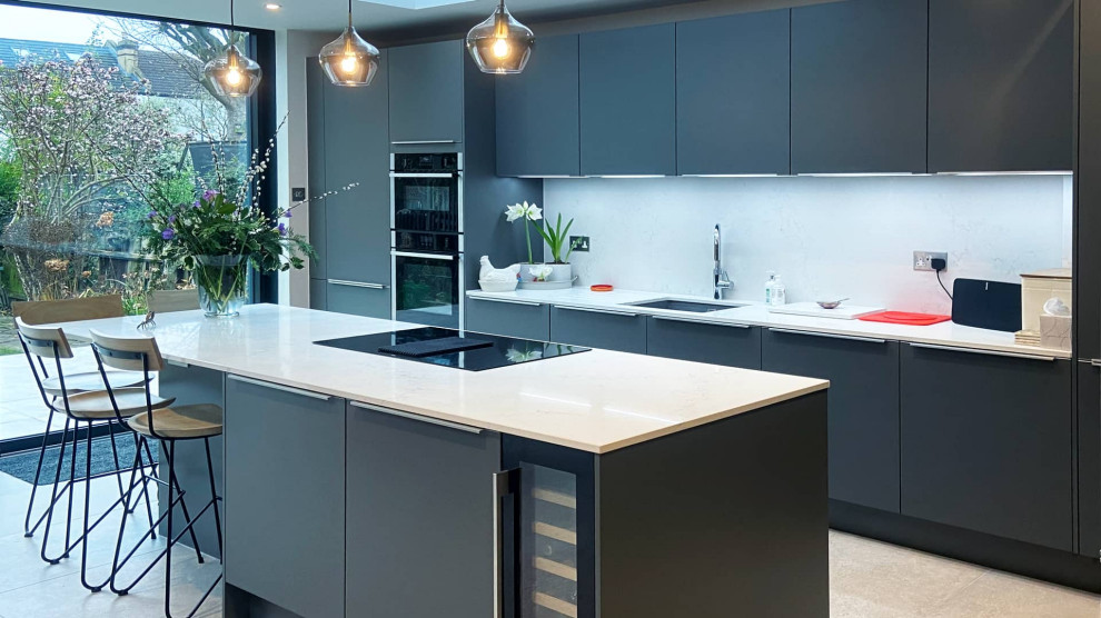 This is an example of an expansive modern grey and white single-wall open plan kitchen in London with a single-bowl sink, flat-panel cabinets, grey cabinets, quartz worktops, white splashback, engineered quartz splashback, black appliances, porcelain flooring, an island, white floors, white worktops and feature lighting.