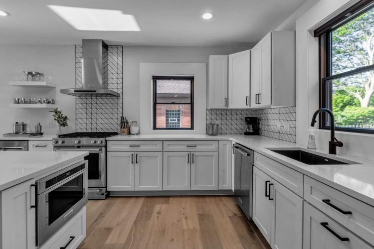 Mid-sized transitional l-shaped light wood floor and brown floor eat-in kitchen photo with an undermount sink, shaker cabinets, white cabinets, quartz countertops, white backsplash, ceramic backsplash, stainless steel appliances, an island and white countertops