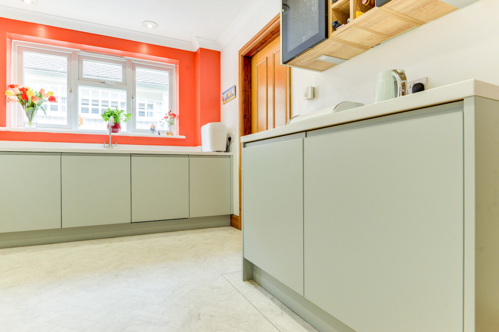 Medium sized beach style grey and white l-shaped enclosed kitchen in Sussex with a built-in sink, flat-panel cabinets, green cabinets, composite countertops, orange splashback, glass sheet splashback, black appliances, lino flooring, a breakfast bar, white floors, white worktops and feature lighting.