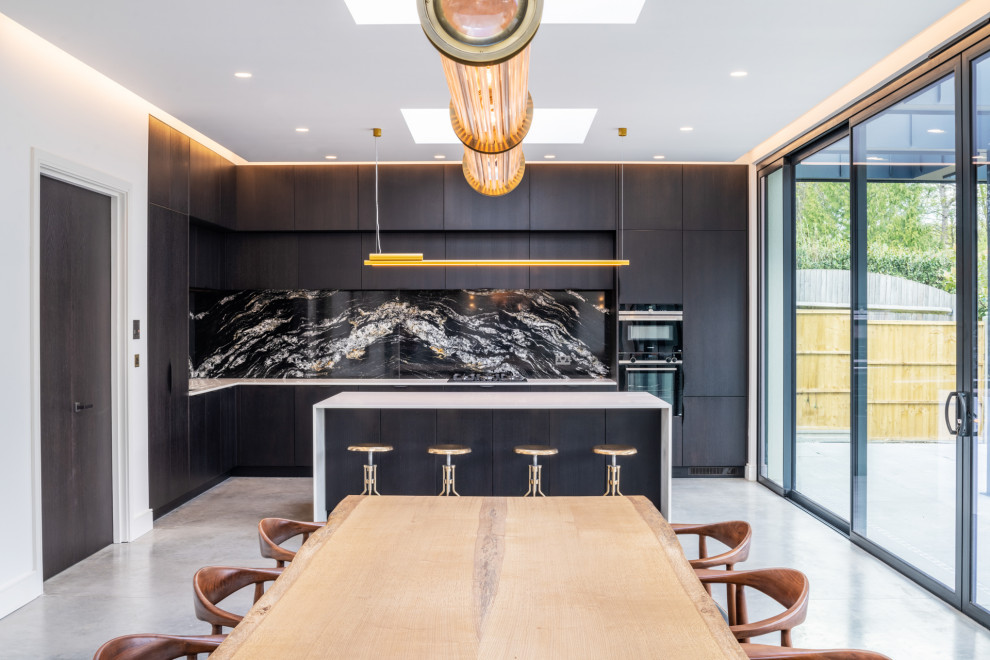 Photo of a contemporary galley kitchen in London with flat-panel cabinets, black cabinets, black splashback, stone slab splashback, black appliances, concrete flooring, an island, grey floors and white worktops.