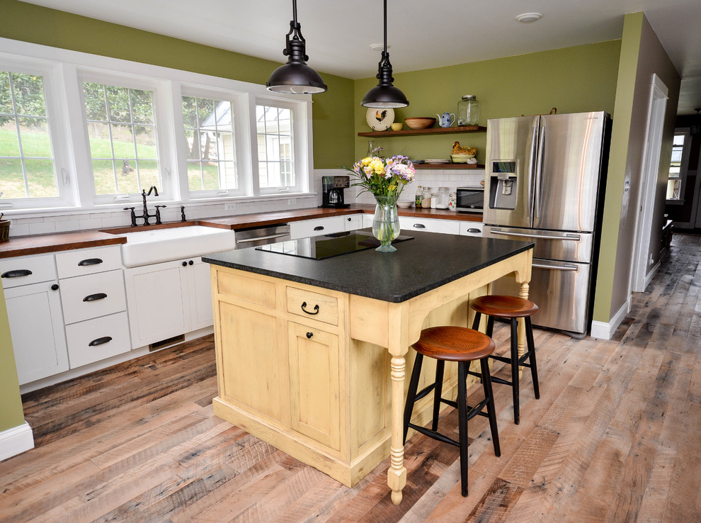 Photo of a mid-sized country l-shaped eat-in kitchen in DC Metro with a farmhouse sink, shaker cabinets, yellow cabinets, granite benchtops, white splashback, subway tile splashback, stainless steel appliances, medium hardwood floors, with island and brown floor.