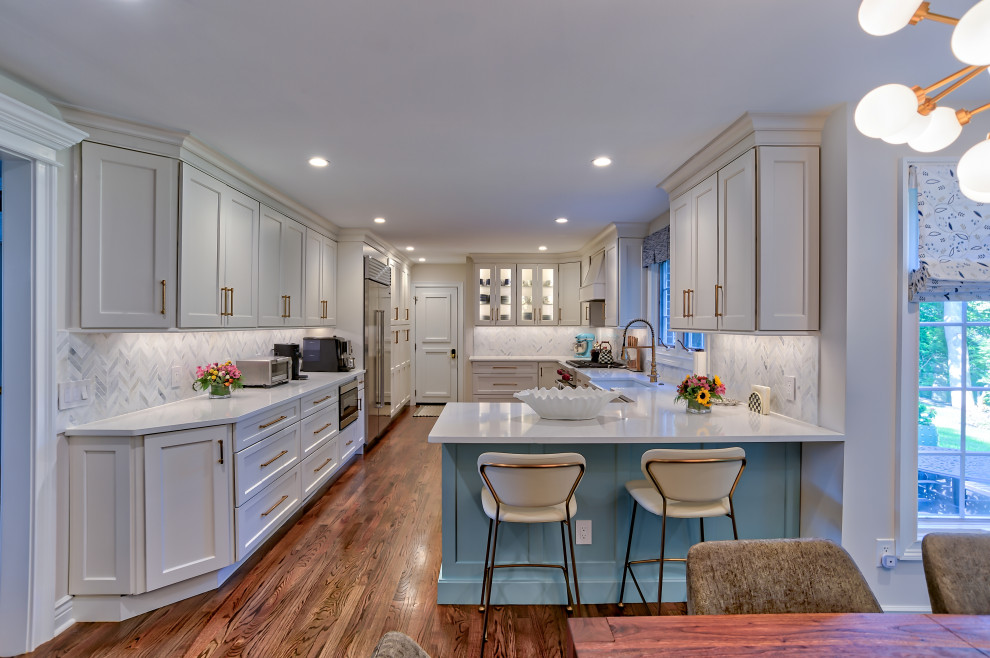 This is an example of a mid-sized transitional u-shaped eat-in kitchen in Philadelphia with a farmhouse sink, recessed-panel cabinets, beige cabinets, quartz benchtops, beige splashback, porcelain splashback, stainless steel appliances, medium hardwood floors, a peninsula, brown floor and white benchtop.