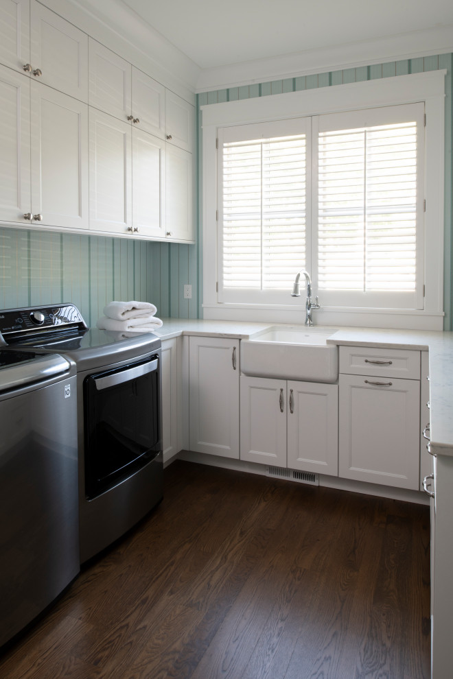 This is an example of a mid-sized beach style u-shaped dedicated laundry room in Minneapolis with a farmhouse sink, recessed-panel cabinets, white cabinets, marble benchtops, multi-coloured walls, dark hardwood floors, a side-by-side washer and dryer, brown floor, white benchtop and wallpaper.