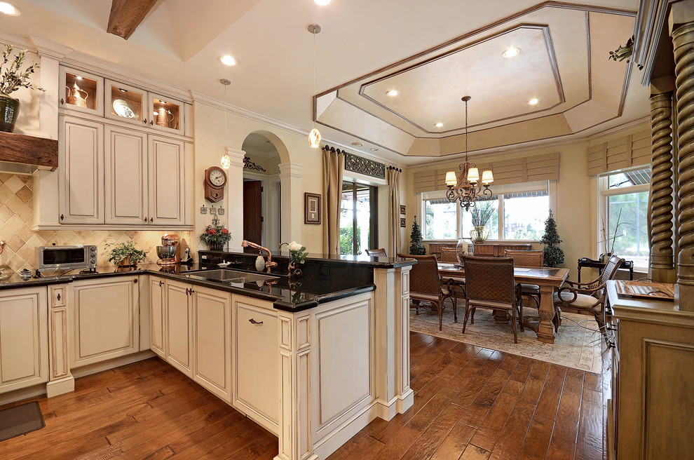 Photo of a large mediterranean galley eat-in kitchen in San Francisco with a drop-in sink, raised-panel cabinets, white cabinets, marble benchtops, beige splashback, stone tile splashback, stainless steel appliances, dark hardwood floors, with island, brown floor and black benchtop.