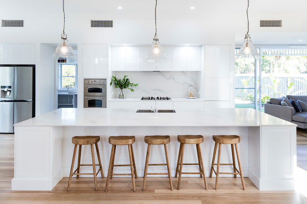 This is an example of a beach style open plan kitchen in Sunshine Coast with an undermount sink, shaker cabinets, white cabinets, white splashback, stainless steel appliances, light hardwood floors, with island and beige floor.