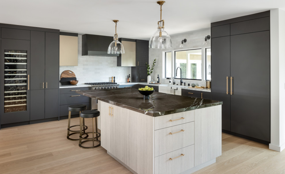 Large contemporary l-shaped eat-in kitchen in Los Angeles with a farmhouse sink, flat-panel cabinets, black cabinets, marble benchtops, white splashback, ceramic splashback, stainless steel appliances, light hardwood floors, with island and multi-coloured benchtop.