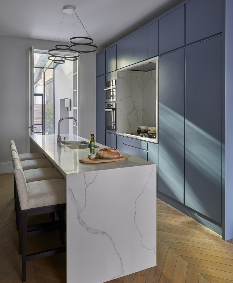 Design ideas for a large contemporary kitchen in London with a drop-in sink, flat-panel cabinets, blue cabinets, marble benchtops, white splashback, marble splashback, black appliances, bamboo floors, with island, beige floor and white benchtop.