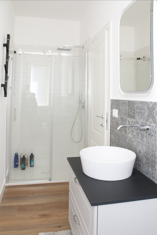 This is an example of a mid-sized contemporary 3/4 bathroom in Other with raised-panel cabinets, grey cabinets, an alcove shower, a wall-mount toilet, gray tile, cement tile, white walls, laminate floors, a vessel sink, laminate benchtops, a sliding shower screen, black benchtops, a single vanity and a freestanding vanity.