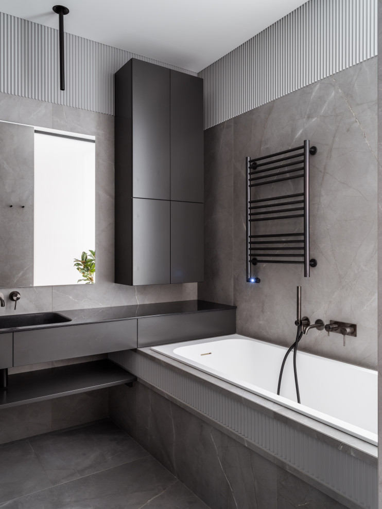 Inredning av ett modernt mellanstort grå grått badrum med dusch, med släta luckor, grå skåp, ett undermonterat badkar, en vägghängd toalettstol, grå kakel, porslinskakel, grå väggar, klinkergolv i porslin, ett integrerad handfat, bänkskiva i akrylsten, grått golv och dusch med gångjärnsdörr