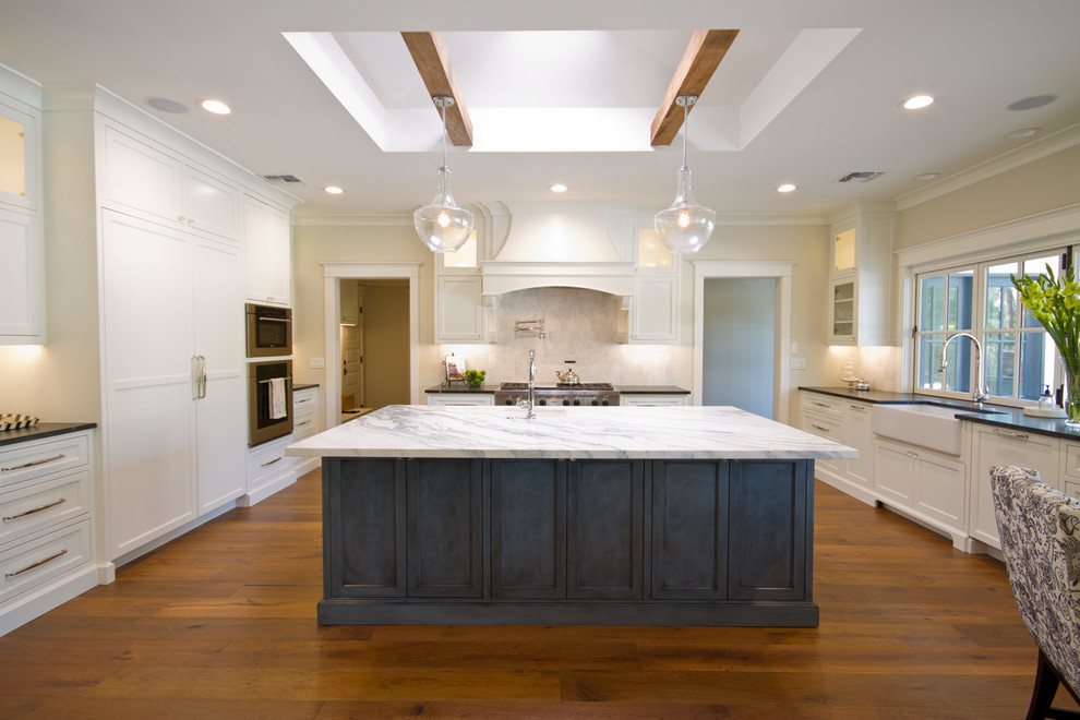 Large transitional u-shaped medium tone wood floor eat-in kitchen photo in Phoenix with a farmhouse sink, recessed-panel cabinets, white cabinets, marble countertops, white backsplash, stone tile backsplash, stainless steel appliances and an island
