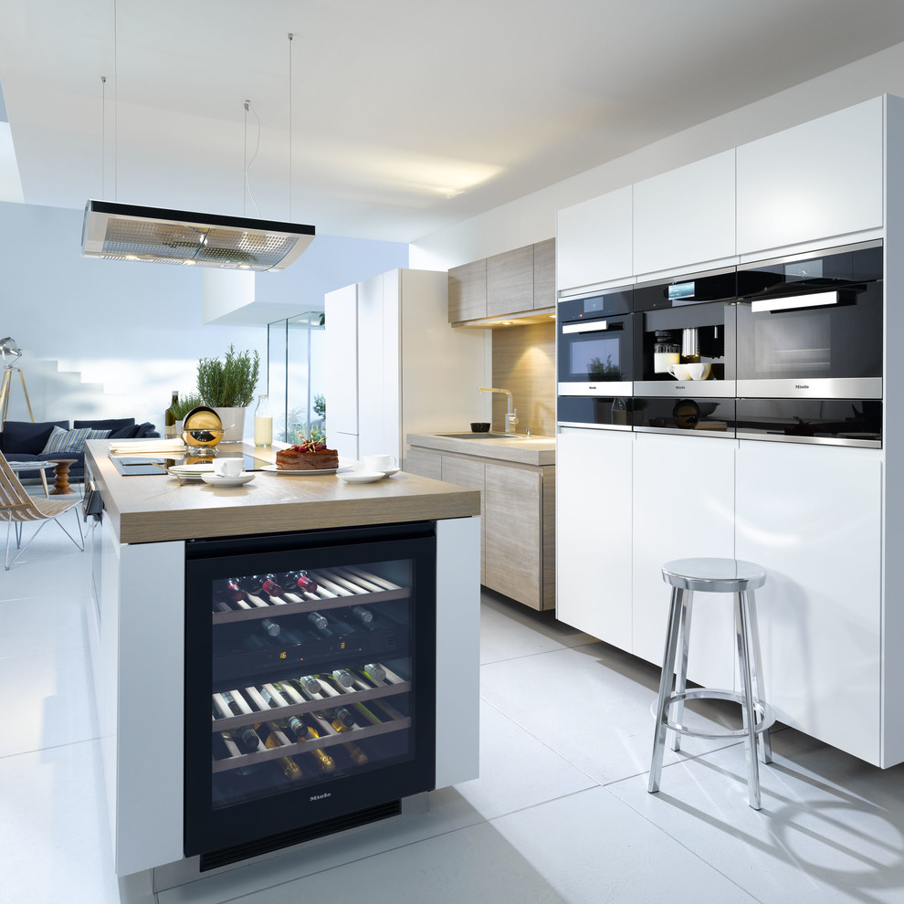 Inspiration for a large contemporary galley open plan kitchen in Oxfordshire with white cabinets, wood benchtops, brown splashback, black appliances, ceramic floors and with island.